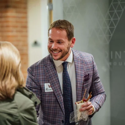 Man in a plaid suit with a name tag smiles while holding food, speaking to a woman with blonde hair at an indoor event.