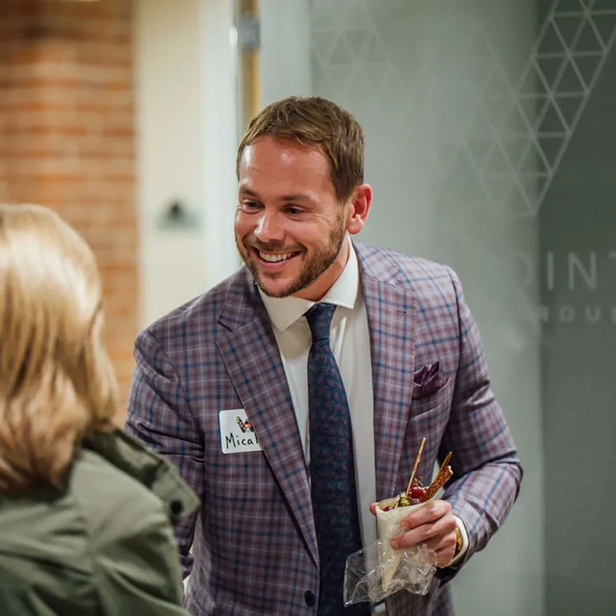 Man in a plaid suit with a name tag smiles while holding food, speaking to a woman with blonde hair at an indoor event.