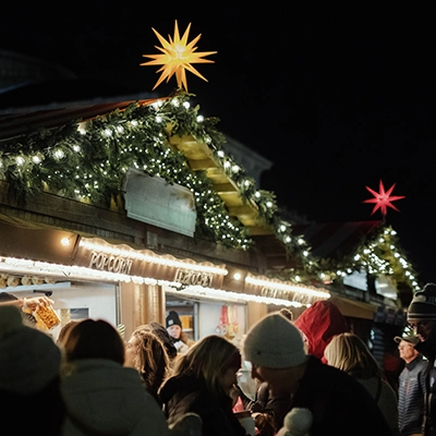 Crowd gathers at a festive outdoor market stall decorated with garlands, string lights, and illuminated star ornaments at night.