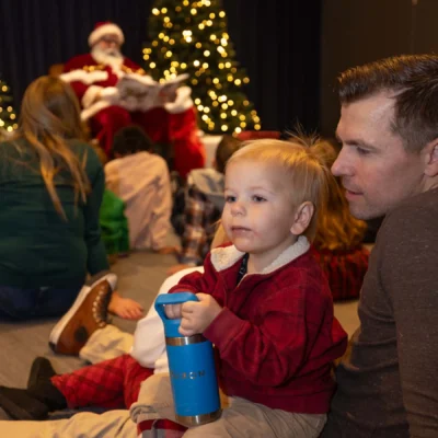 A young child holding a blue cup sits on an adult’s lap, with people gathered in front of a Santa Claus figure and lit Christmas trees in the background.