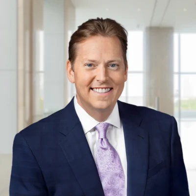 A man in a dark suit with a light purple patterned tie smiles at the camera in a modern office setting with large windows.