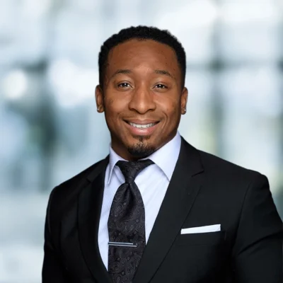 A man wearing a black suit, white shirt, and patterned tie, standing in front of a blurred background, smiling at the camera.