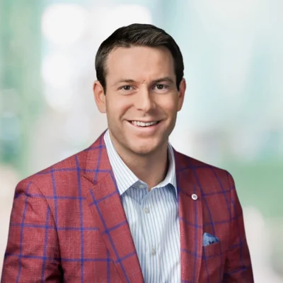 A man with short dark hair is wearing a red plaid blazer over a light blue striped shirt, smiling at the camera with a blurred background.