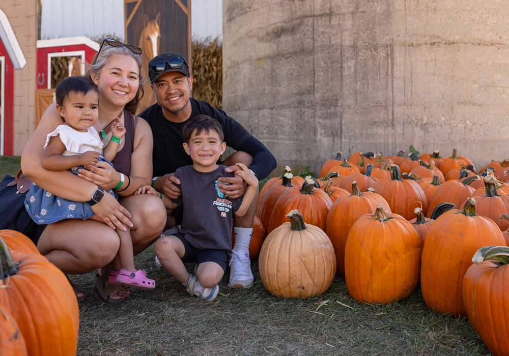 A family of four poses and smiles next to a group of pumpkins outdoors at what appears to be a pumpkin patch or fall festival.