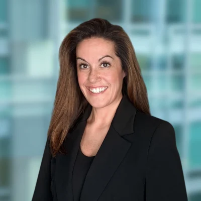 Woman with long brown hair in a black blazer smiling, standing in front of a blurred office building background.