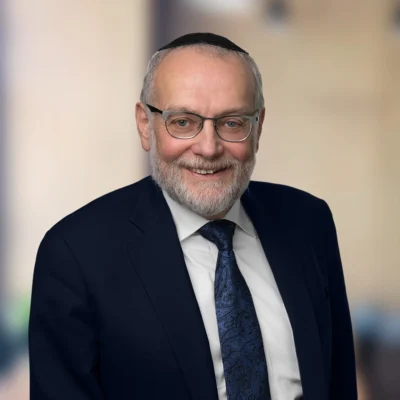 An older man with glasses, a beard, and a dark suit with a blue patterned tie smiles at the camera against a blurred background.