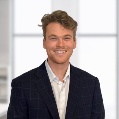 A young man with wavy hair wearing a dark checked blazer and a white shirt stands indoors, smiling at the camera.