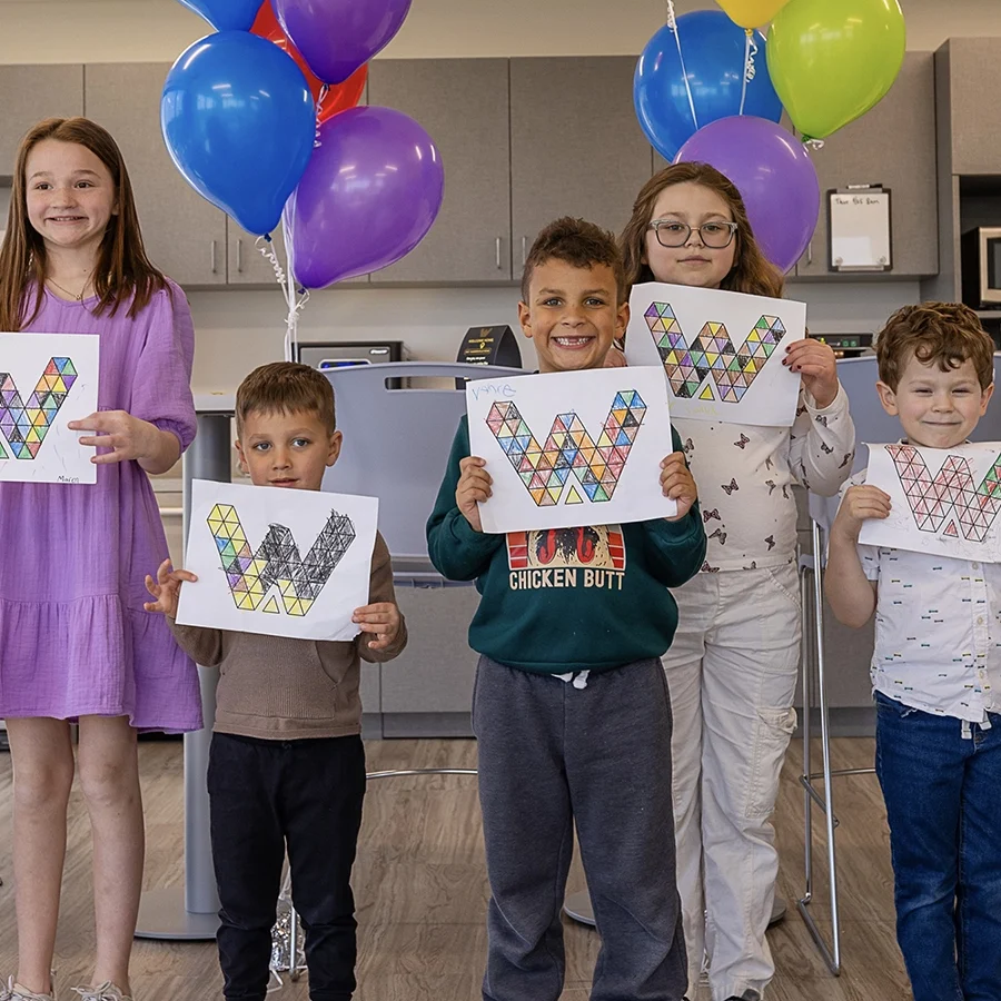 Five children stand indoors holding up colored letter "W" drawings, with colorful balloons in the background.