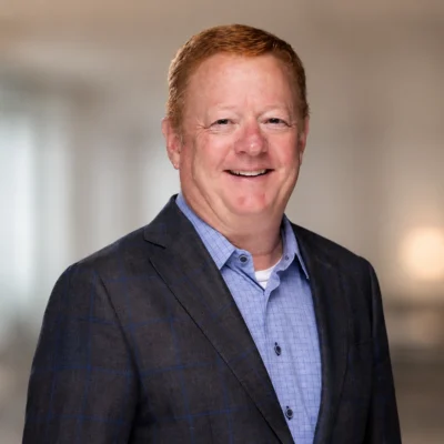 A middle-aged man with short red hair, wearing a blue plaid suit jacket over a light blue dress shirt, smiles at the camera in a softly blurred indoor setting.