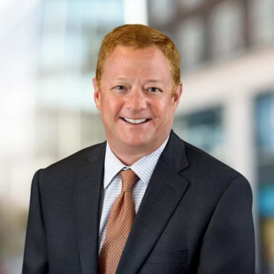 A man with short red hair in a suit and tie smiles at the camera. The background is a blurred cityscape.