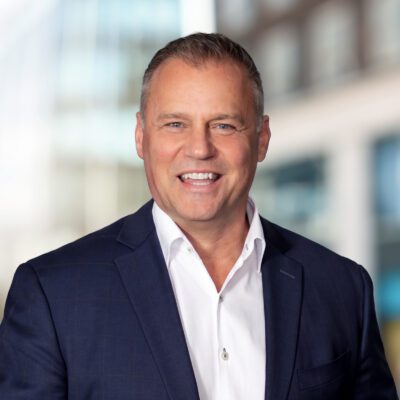 A middle-aged man in a navy suit and white shirt smiles, standing in front of a blurred urban background.