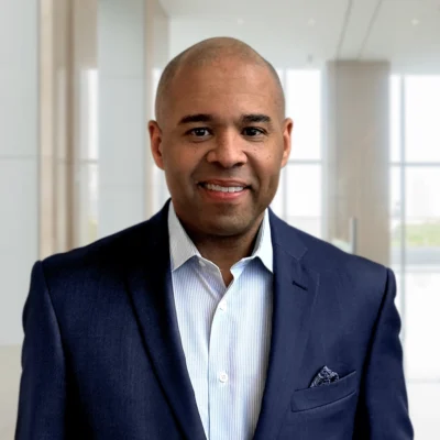 A man in a dark blue suit and striped shirt stands indoors in a modern office setting, smiling at the camera.