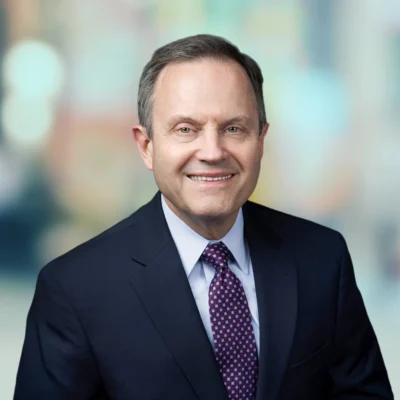 A middle-aged man in a dark suit and patterned tie smiles at the camera against a blurred background.