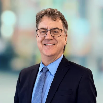 A man wearing glasses, a dark suit, blue shirt, and tie is smiling at the camera with a blurred background behind him.