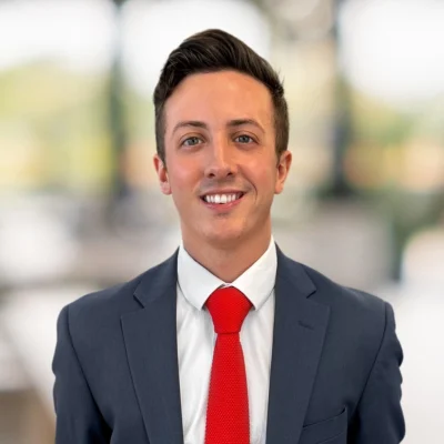 A young man in a suit and red tie smiles at the camera with a blurred office background.