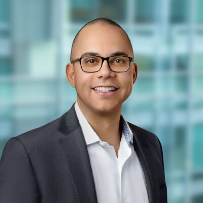 A man in a suit and glasses smiles at the camera in front of a blurred, modern office background.