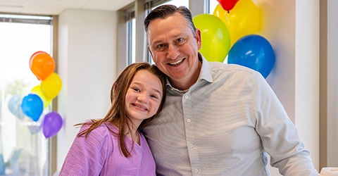 A man and a young girl smile while standing together indoors with colorful balloons in the background.