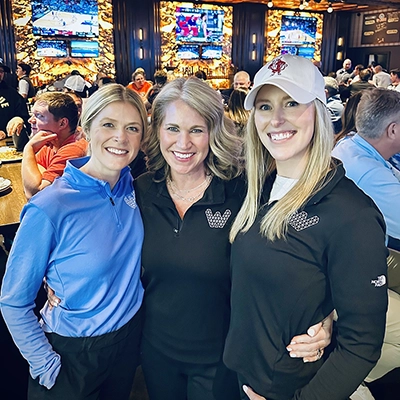 Three women smile and pose together in a busy sports bar, with multiple TV screens and people in the background.