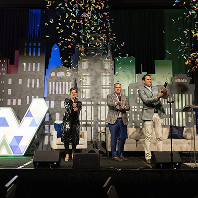 Three people on stage celebrate by popping confetti cannons in front of a cityscape backdrop with a large geometric "W" and a couch behind them.