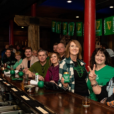 A group of people wearing green sit and stand at a bar decorated with Guinness banners. The atmosphere suggests a St. Patrick's Day celebration.