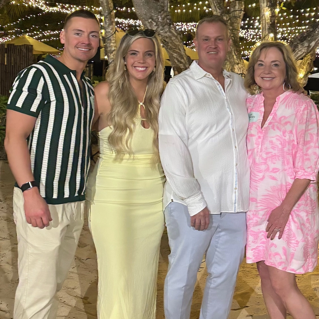 Four adults stand together outdoors at night under string lights, dressed in light-colored, summery clothing, smiling at the camera.
