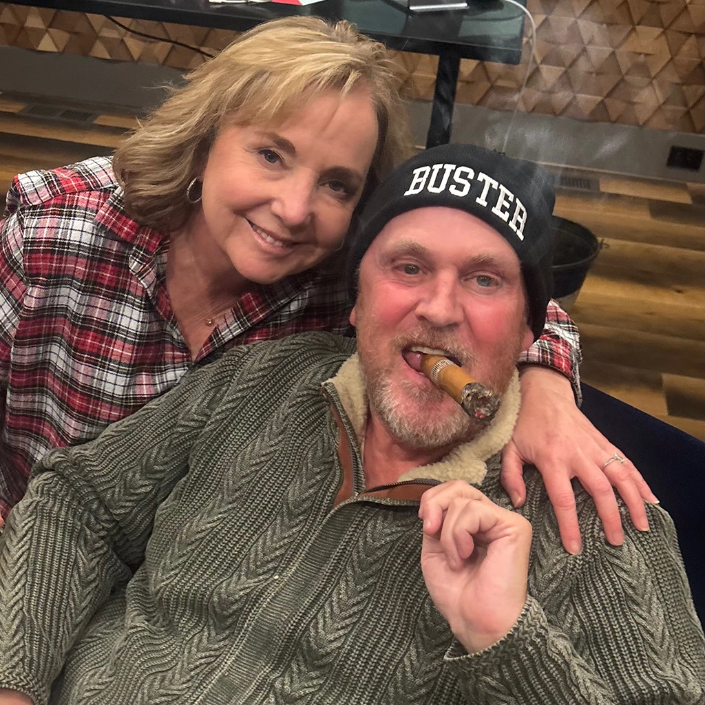 A woman in a plaid shirt smiles with her arm around a man in a knit sweater and "BUSTER" beanie, who is holding a cigar and looking at the camera indoors.