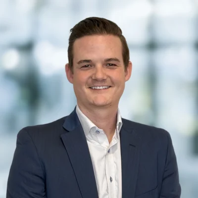 A man in a suit jacket and white shirt is smiling in front of a blurred background.