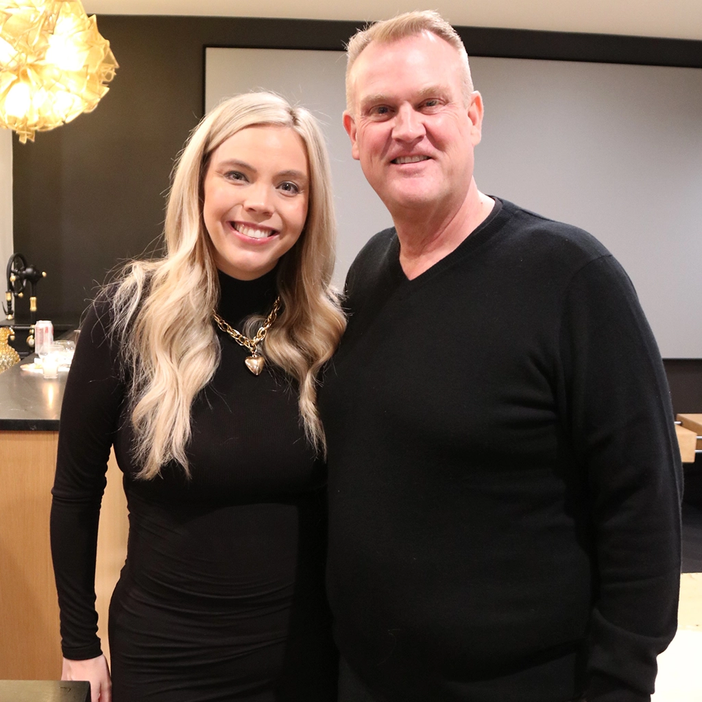A woman and a man stand side by side smiling at the camera indoors, both wearing black tops.