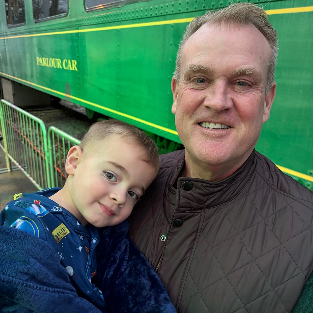 An adult and a child stand together in front of a green train car labeled "Parlour Car." The child is wrapped in a blue blanket and leaning on the adult.