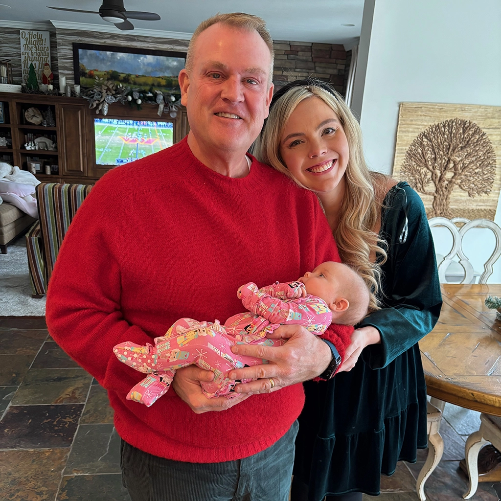 A man in a red sweater holds a baby in pink pajamas, standing next to a smiling woman in a green dress inside a living room.