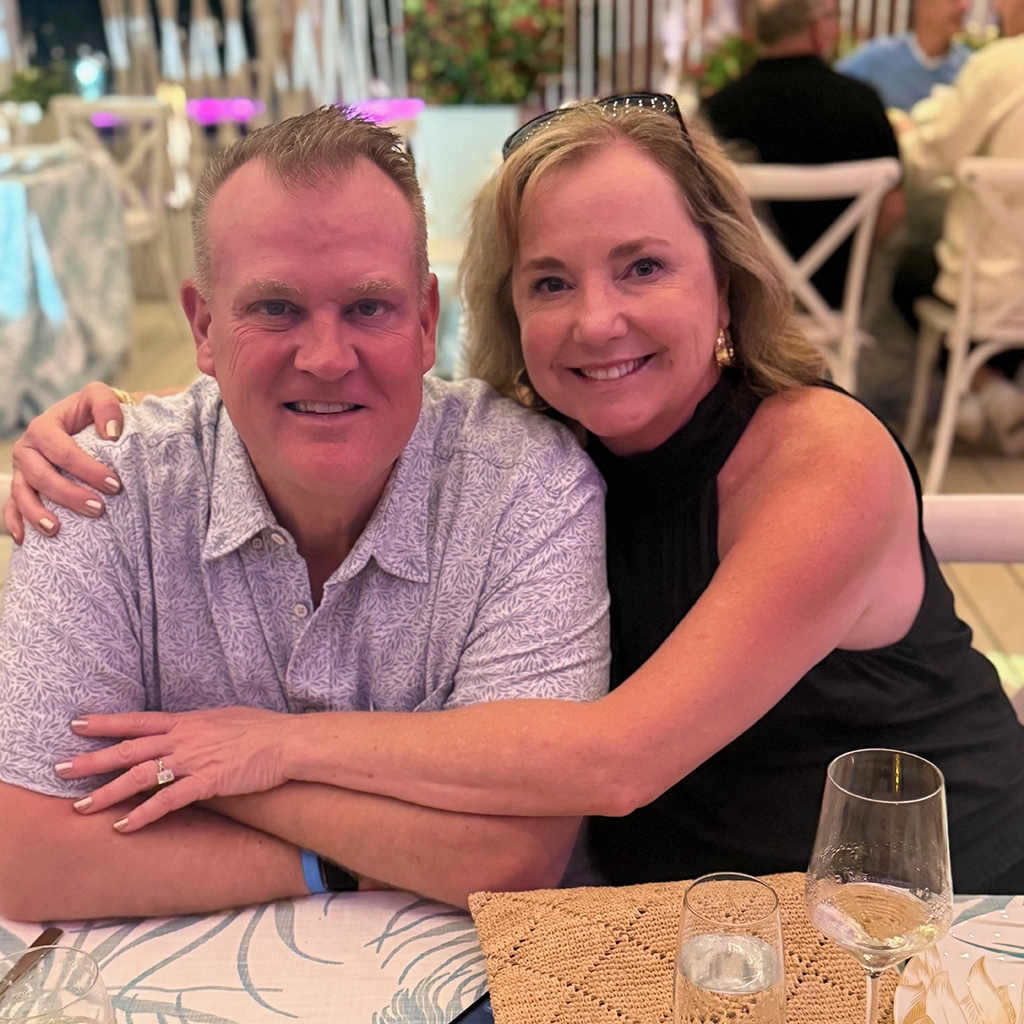 A man and woman sit closely together at a restaurant table, smiling at the camera. The table has drinks, and other diners are visible in the background.