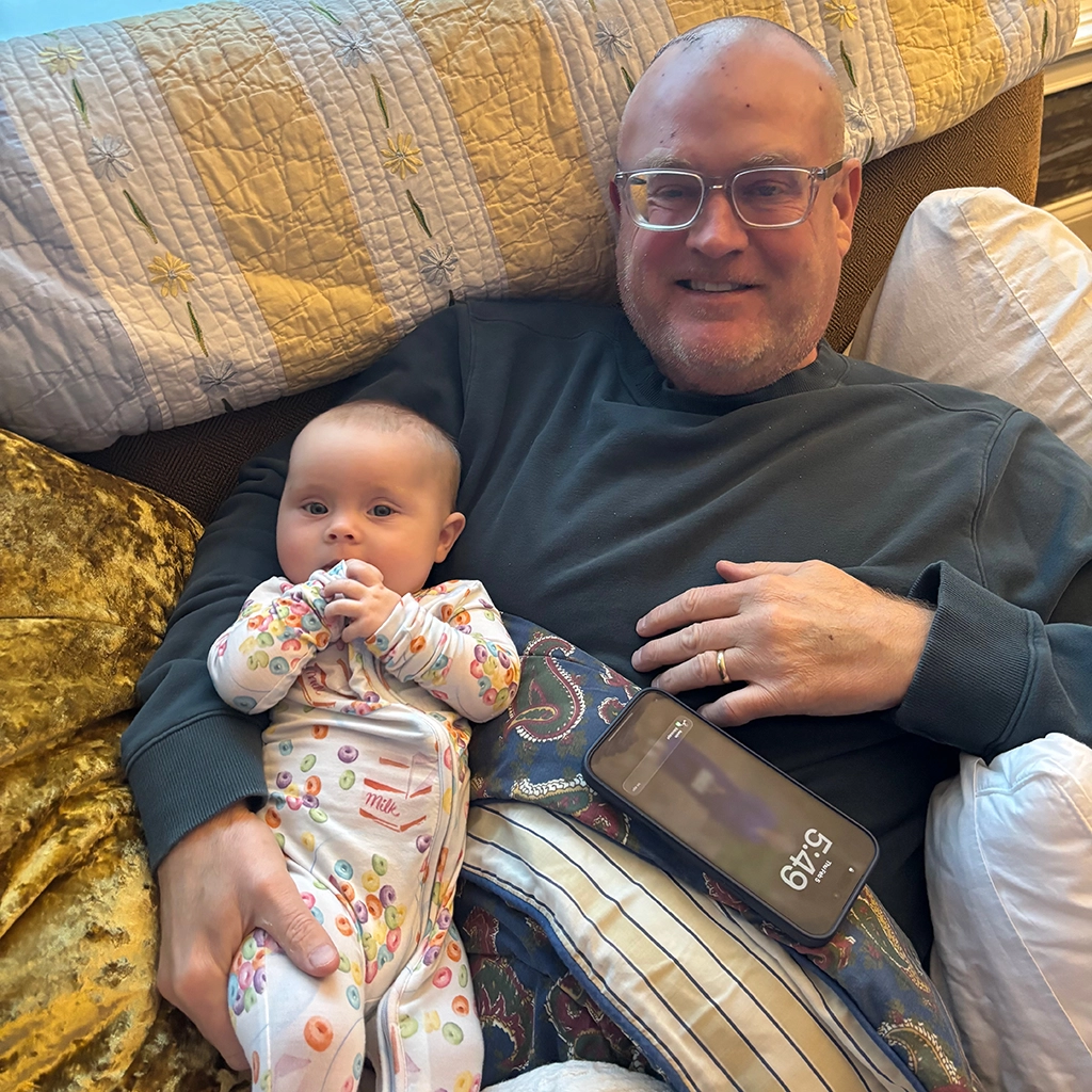 A man wearing glasses reclines on a couch holding a baby in patterned pajamas, with a smartphone resting on his lap displaying the time as 5:49.