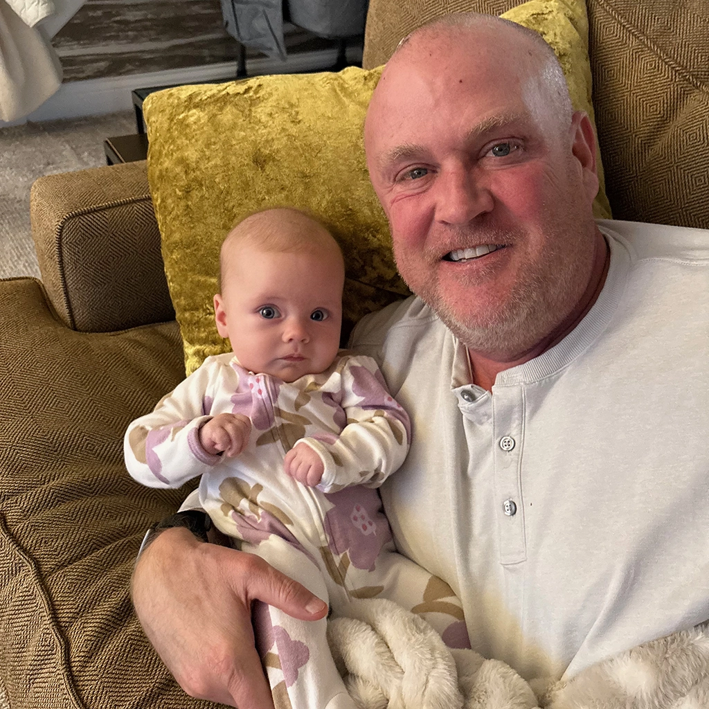 A man sits on a couch holding a baby dressed in a patterned onesie; both are looking at the camera and smiling.