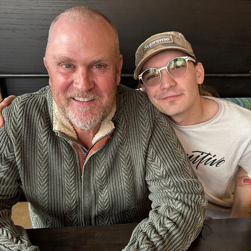 Two men sit closely together, smiling at the camera. One wears a knit sweater, and the other wears glasses, a cap, and a t-shirt.