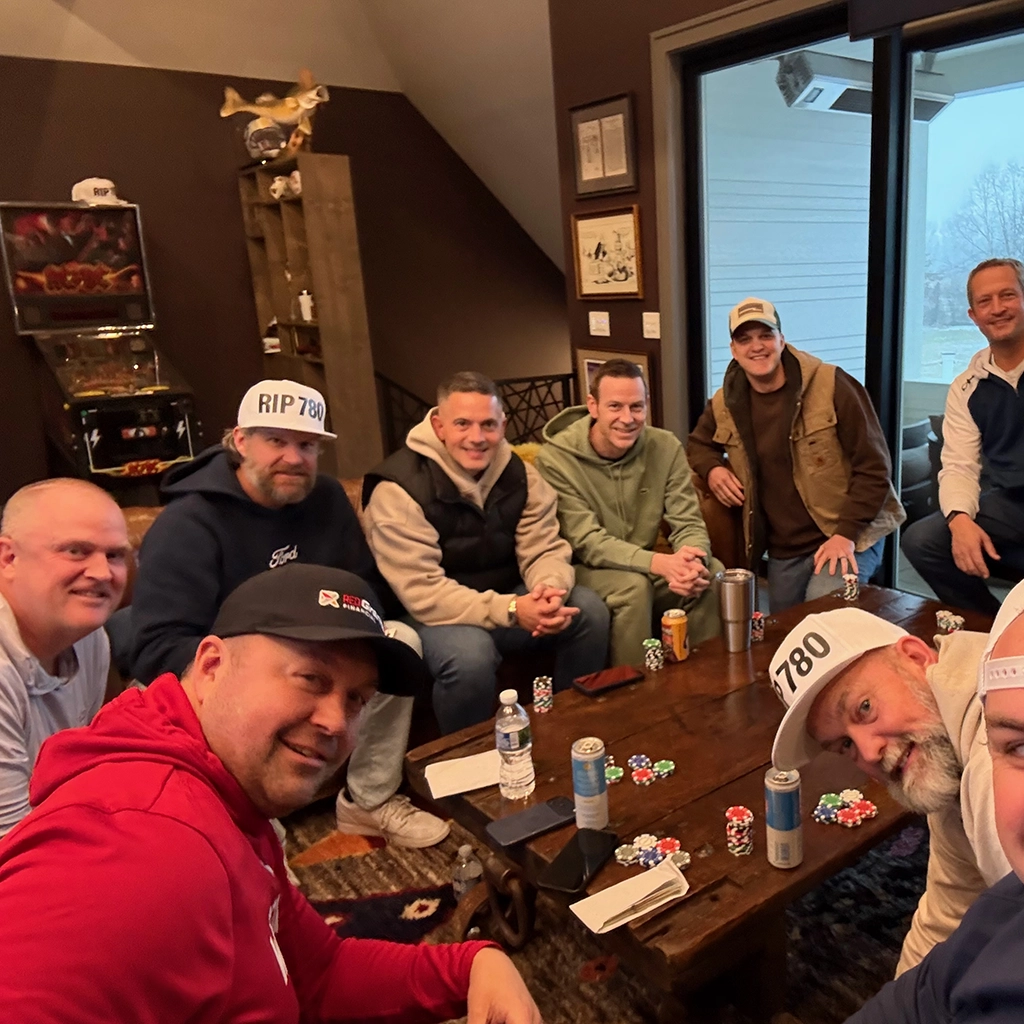 Nine men sit around a coffee table with poker chips, playing cards, and drinks in a casual indoor setting. Some wear hats, and there is a pinball machine in the background.