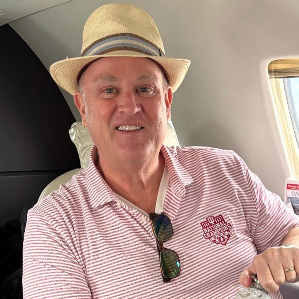 A man wearing a striped polo shirt and straw hat sits in an airplane seat, smiling at the camera. Sunglasses hang from his shirt collar.