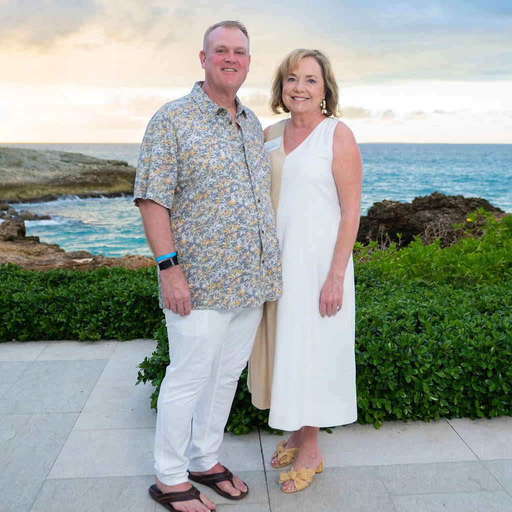 A man and woman stand together outside near green bushes with the ocean and a cloudy sunset in the background.