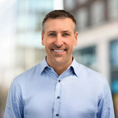 Smiling man wearing a light blue button-up shirt standing outdoors with blurred buildings in the background.