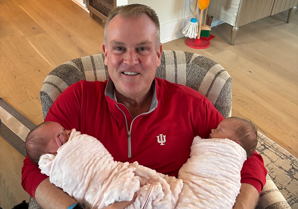 A man in a red Indiana University pullover sits in a chair, smiling and holding two swaddled infants, one in each arm.