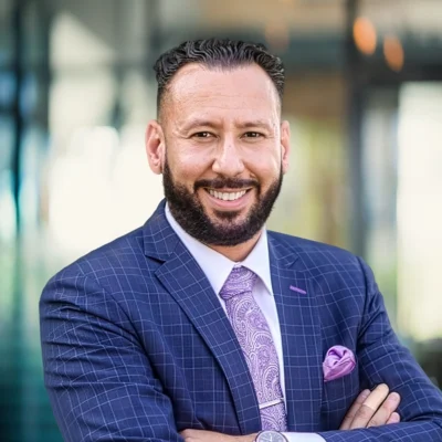 A man in a blue checkered suit and patterned tie smiles with arms crossed, standing in front of a blurred office building background.