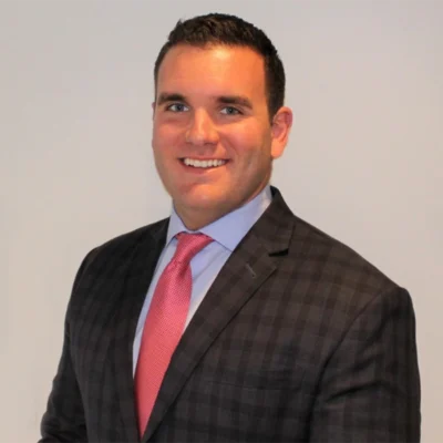 A man in a dark plaid suit, light blue shirt, and pink tie smiles at the camera against a plain light-colored background.