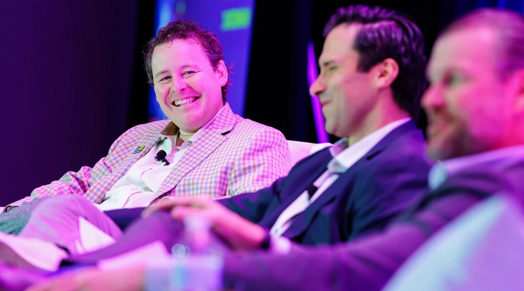 Three men in business attire sit and converse on a panel discussion stage, one of them smiling broadly, with microphones clipped to their jackets.