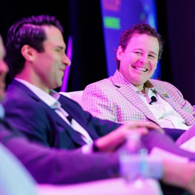 Three men in business attire sit on a panel discussion stage, conversing and smiling under colorful lighting.