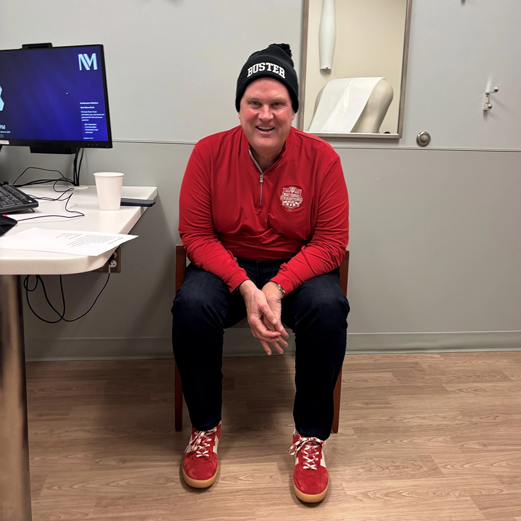A person wearing a red shirt, dark pants, red shoes, and a black beanie sits on a chair in a medical office, smiling at the camera.