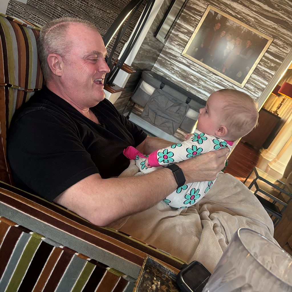 A man sits in a striped armchair holding a baby dressed in a floral onesie; they are facing each other and smiling in a warmly lit living room.