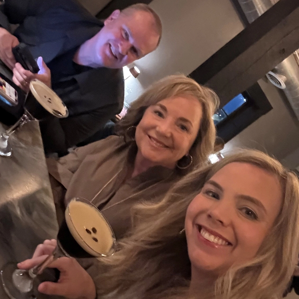 Three people sit together at a bar table with espresso martinis, smiling at the camera in a dimly lit setting.