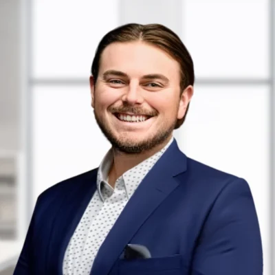 A man with brown hair and a beard, wearing a navy blue suit and white patterned shirt, smiles while standing in a bright office setting.