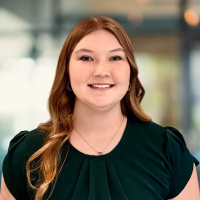 Woman with long, wavy auburn hair wearing a dark green blouse and necklace, smiling, in an indoor setting with blurred background.