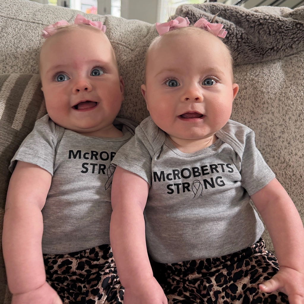 Two babies with pink bows wearing matching "McROBERTS STRONG" shirts and leopard print pants sit side by side on a patterned couch.