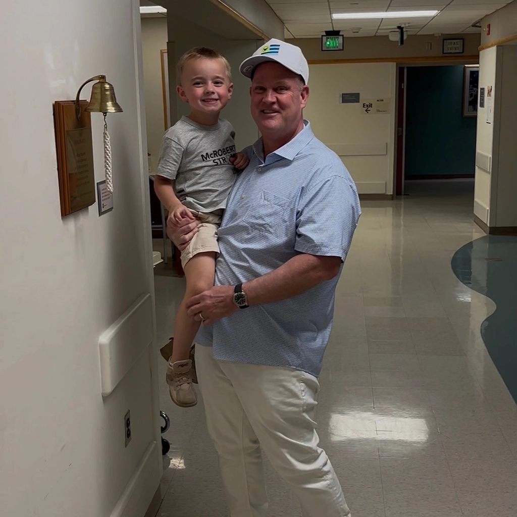 An adult wearing a cap holds a smiling child in a hallway next to a brass bell mounted on the wall.